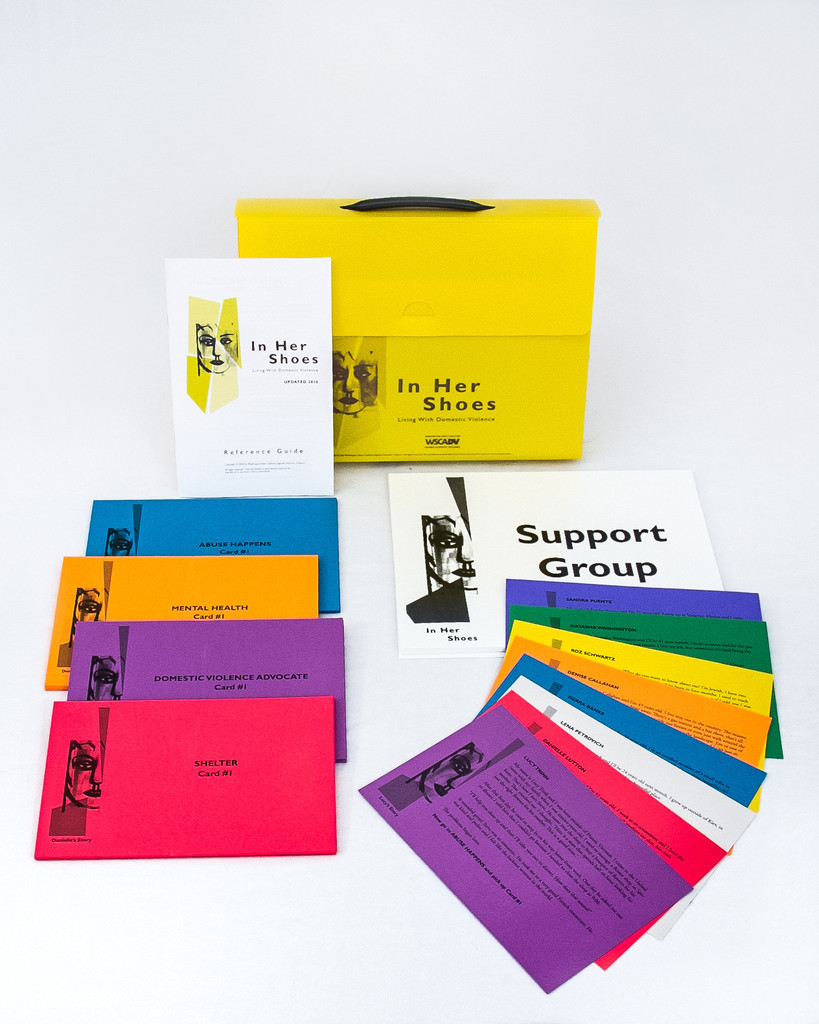 Picture of the original In Her Shoes Training Kit. The picture shows some pieces from the kit laid out on a table including multicolored cards, an instruction booklet, and a station label.  