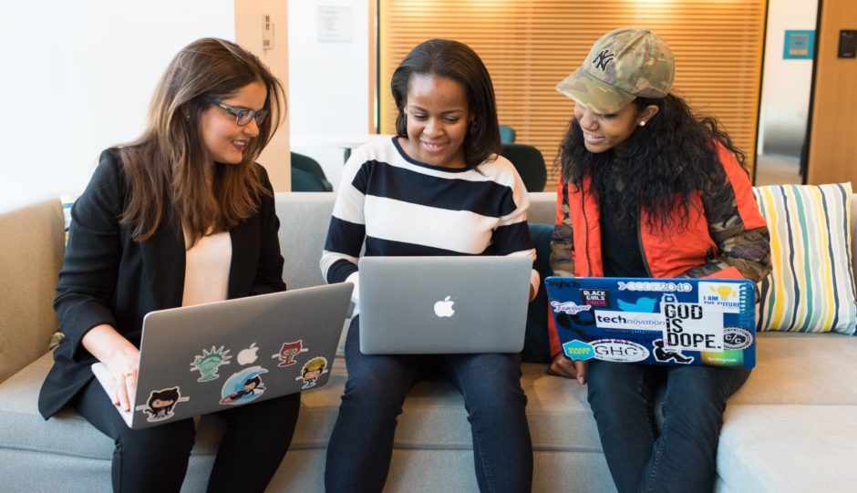 Photo of three young women sitting on a couch. Each one has a laptop open on their lap and they are smiling while they look at the screen.