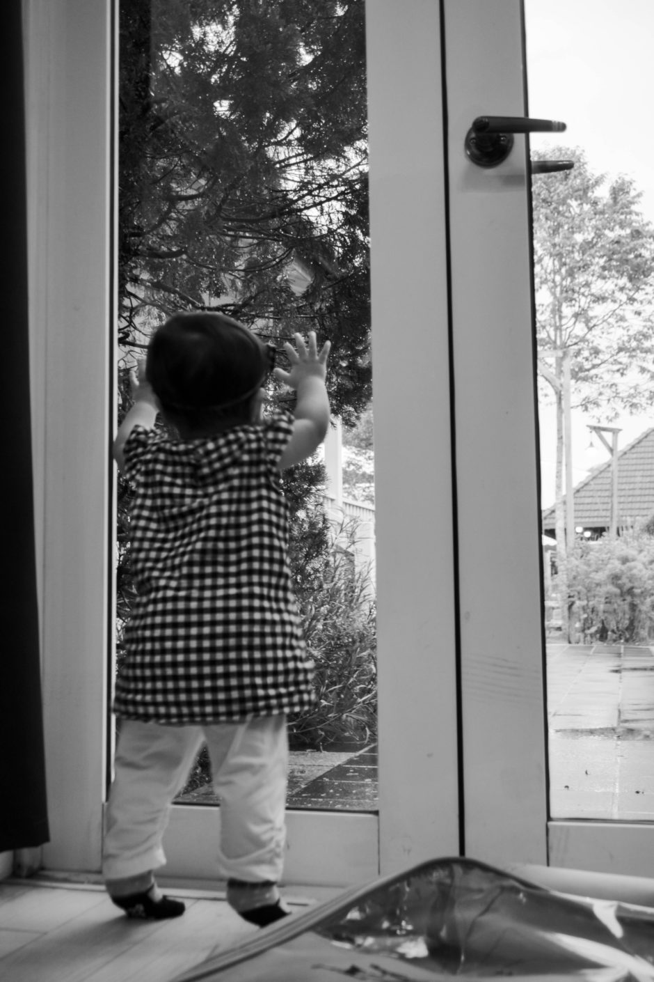 Photo of a toddler standing on their toes looking out a set of glass doors with their hands up on the glass.