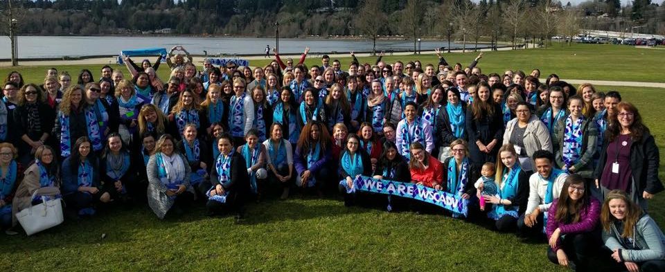 Photo of approximately 100 people outside in front of a lake in Olympia, WA holding scarves that say, "We Are WSCADV."