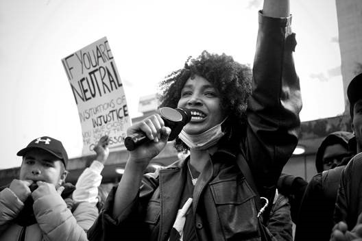 photo of a group of protesters with a young Black woman holding a microphone, smiling, with her arm raised in the air.
