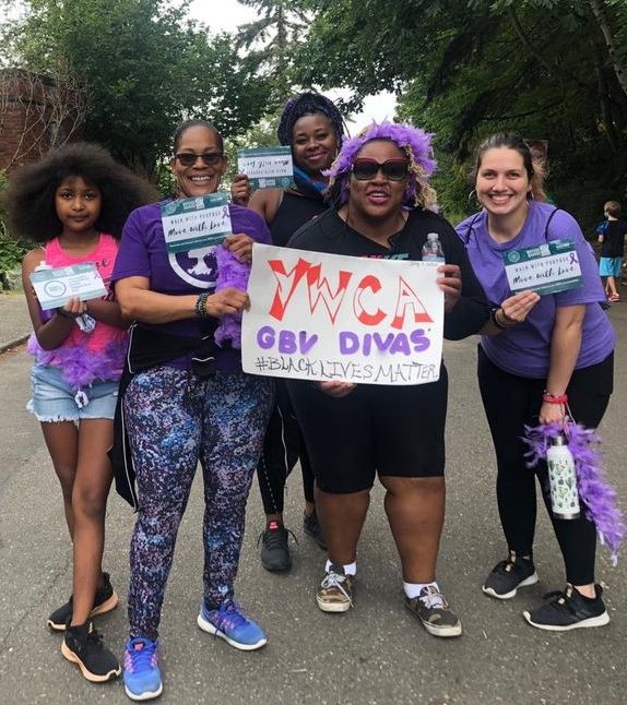 Group of women and children smiling at the camera. They are participating in the 2020 Refuse To Abuse 5K. They are wearing purple and holding a sign that says YWCA GBV Divas #BlackLivesMatter. They are also holding signs that have violence prevention messages on them.