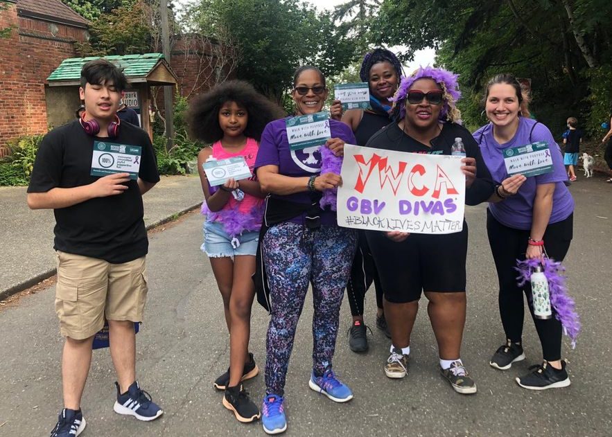 Group of women and children smiling at the camera. They are participating in the 2020 Refuse To Abuse 5K. They are wearing purple and holding a sign that says YWCA GBV Divas #BlackLivesMatter. They are also holding signs that have violence prevention messages on them.