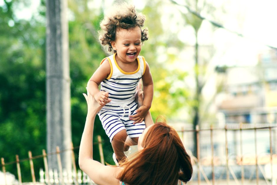 Photo of a woman holding a smiling toddler up in the air.