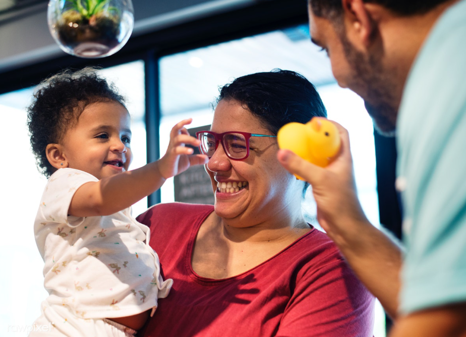 2 adults, 1 offering a rubber duck to a baby, they are all smiling and happy