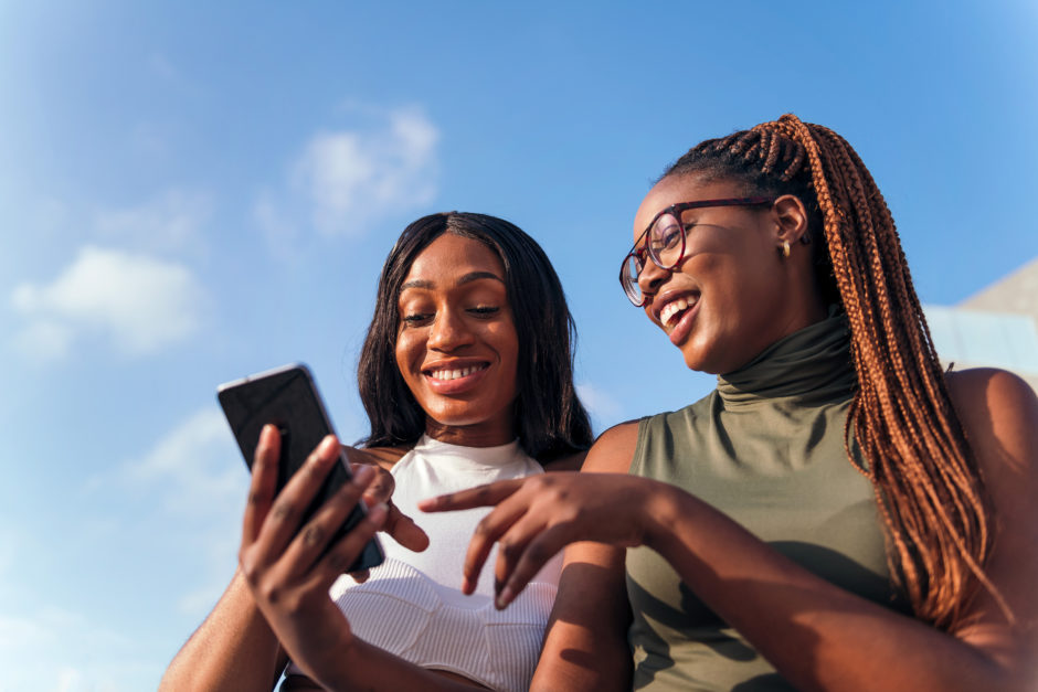 Two Black young women are laughing together and looking at a phone with blue sky in the background.